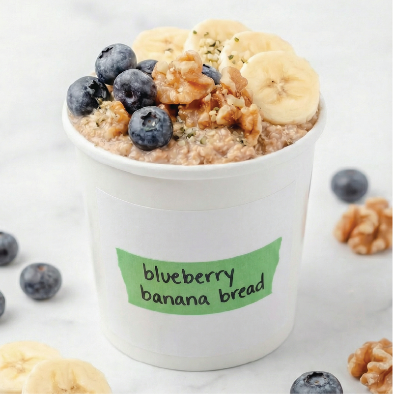 White container of blueberry banana bread flavored oatmeal with blueberries, banana slices, and granola on a white background.