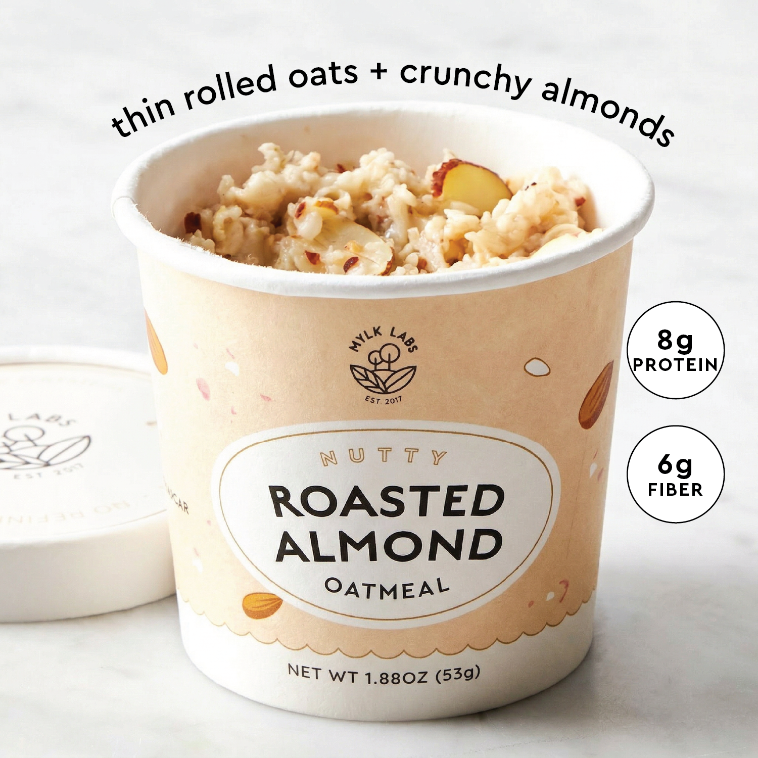 A cup of Mylk Labs Nutty Roasted Almond Oatmeal with thin rolled oats and crunchy almonds visible at the top, shown on a light marble-like surface with the lid beside it. Text overlays read 'thin rolled oats + crunchy almonds' and '8g PROTEIN' and '6g FIBER'.