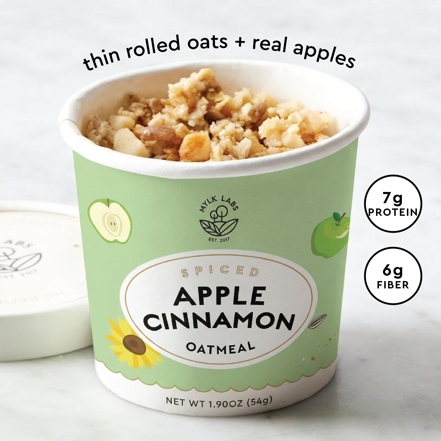 A container of Mylk Labs Spiced Apple Cinnamon Oatmeal, showing the lid off to reveal the oatmeal inside. The label is light green with white and black text, and includes graphic illustrations of an apple, a sunflower, and nutritional badges indicating 7g protein and 6g fiber.