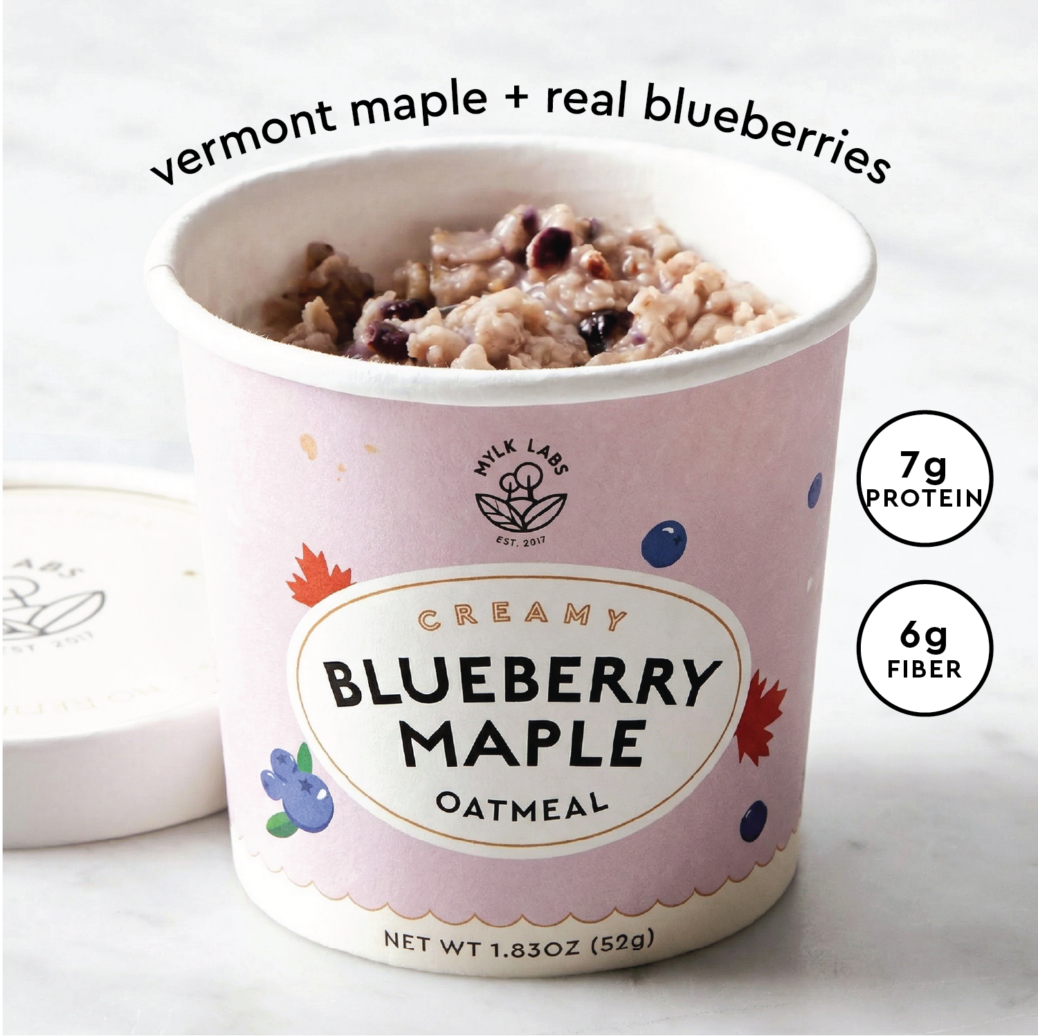 A cup of Mylk Labs Creamy Blueberry Maple Oatmeal with a light pink lid resting beside it on a light marble surface. The cup is filled with oatmeal topped with blueberries, and text on the image reads 'vermont maple + real blueberries' with nutritional callouts '7g PROTEIN' and '6g FIBER'.