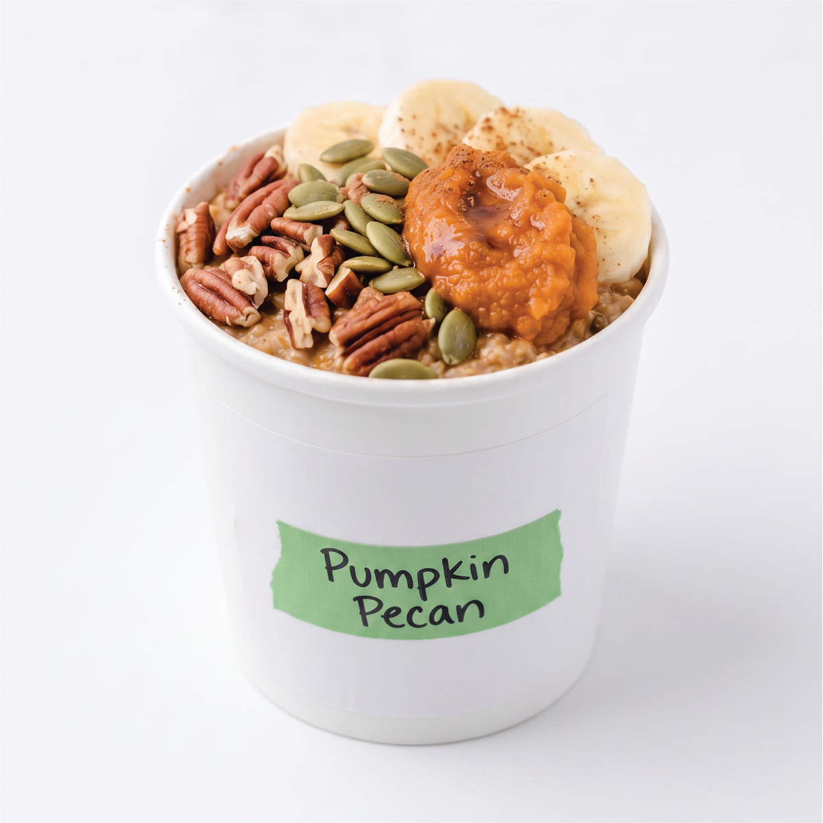 A white paper cup filled with pumpkin pecan oatmeal, topped with sliced bananas, pumpkin pie filling, pecans, and green pumpkin seeds, with a green label reading 'Pumpkin Pecan' on the front.