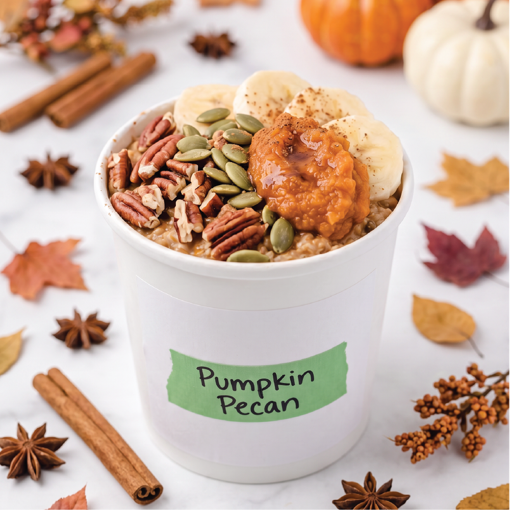 A white cup labeled 'Pumpkin Pecan' filled with pumpkin oatmeal, topped with sliced bananas, green pumpkin seeds, and pecans, styled with autumn leaves, cinnamon sticks, star anise, and small pumpkins on a light marble surface.