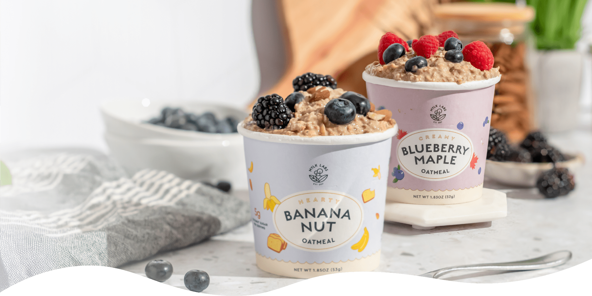 Two containers of oatmeal with toppings on a table, featuring 'Banana Nut' and 'Blueberry Maple' labels.